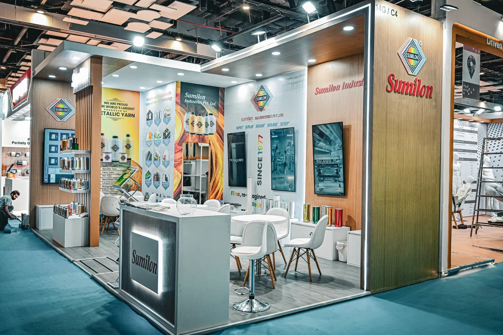 A wide view of the Sumilon stand from the aisle, its corner open on two sides with sample boards, a counter and seating inside.