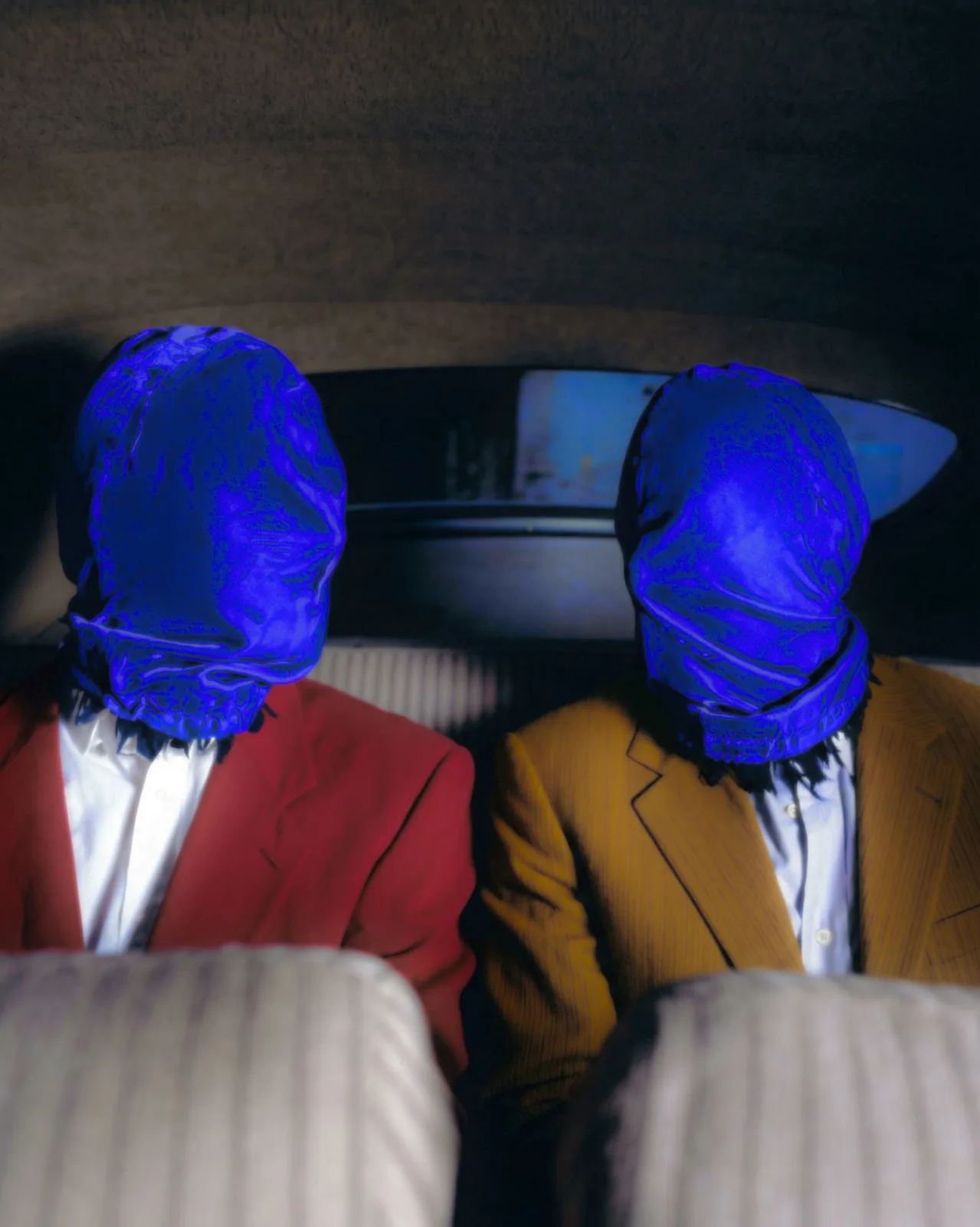 The same two men in the back of a car, shot close, the blue cloth over their faces catching the light against a red and an ochre jacket.