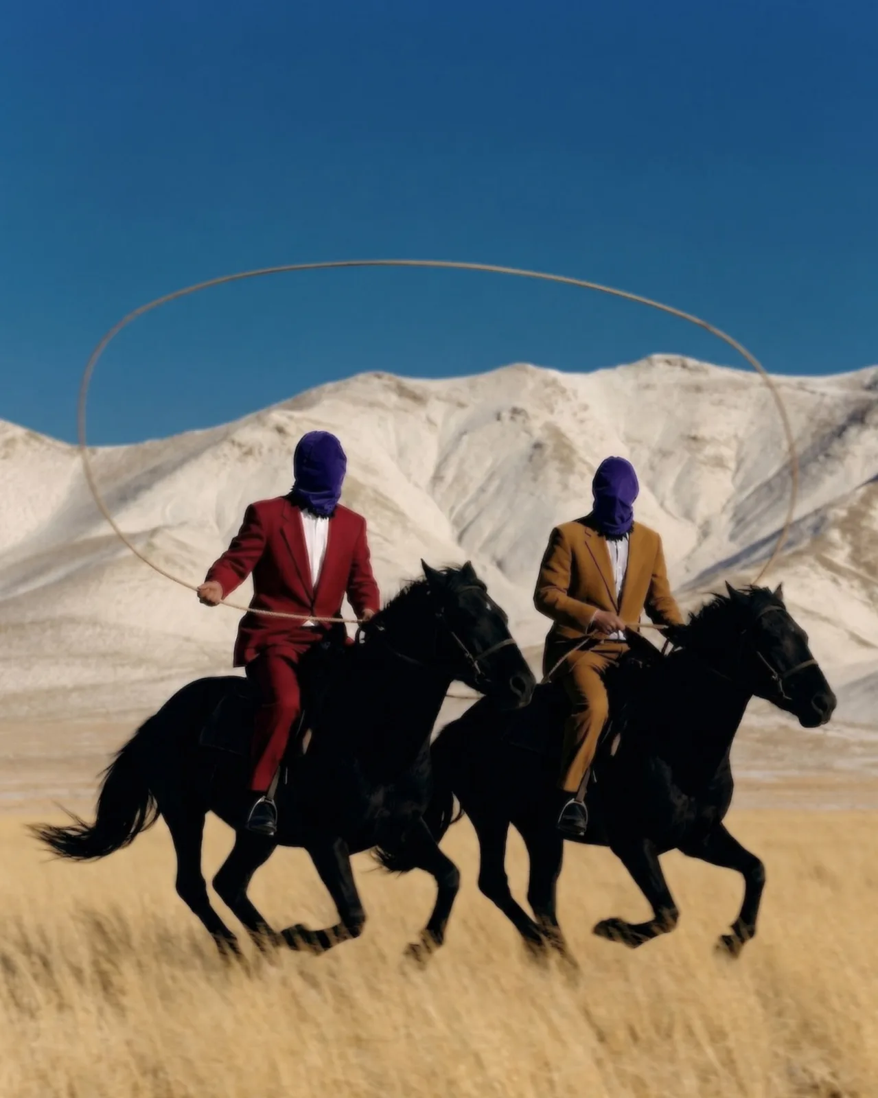 The two men riding black horses at a gallop across dry grassland below white hills, one swinging a rope in a wide loop against a clear blue sky.