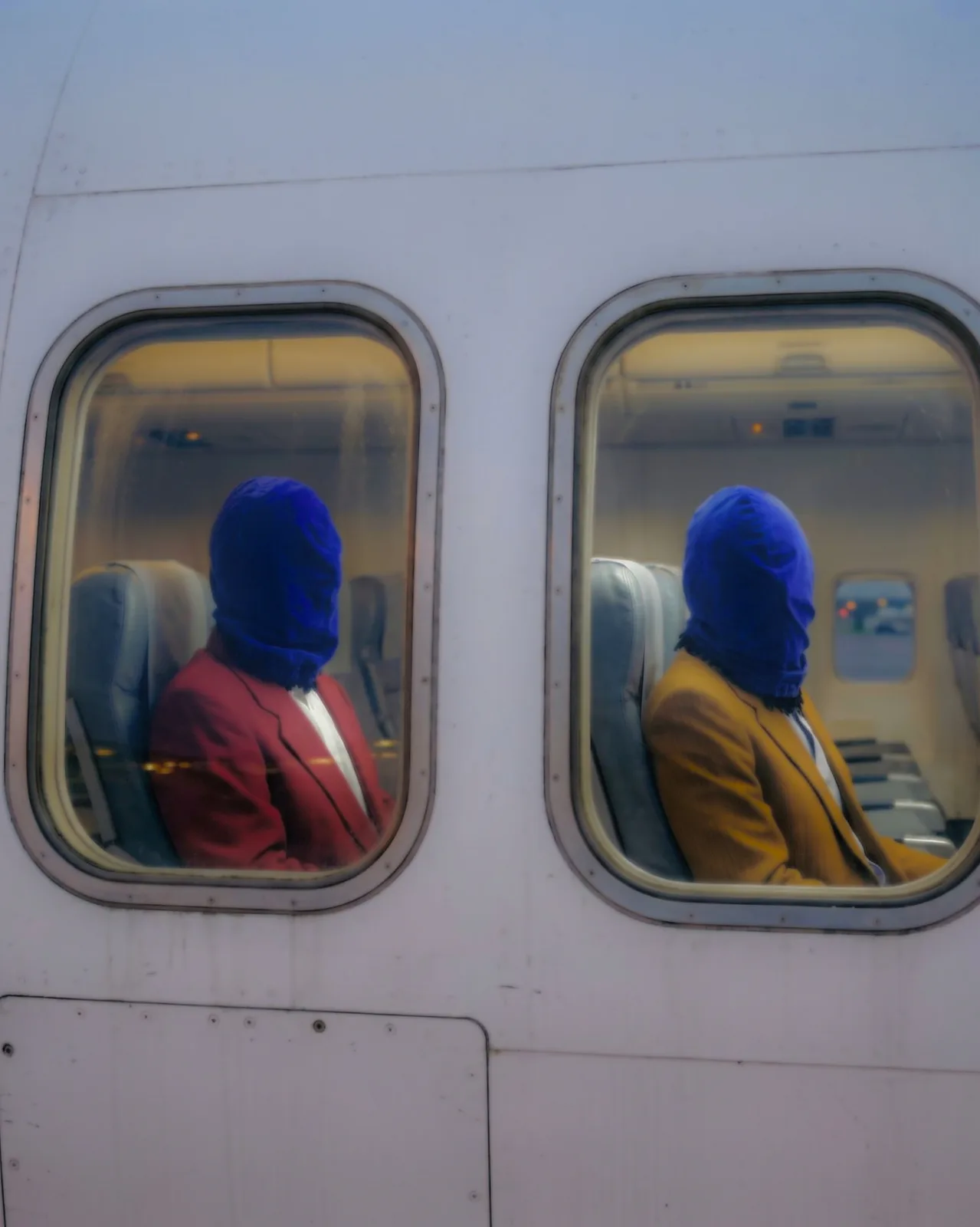 The two men seen through two windows of a parked aircraft, one in red and one in ochre, faces wrapped in blue, waiting in the cabin.