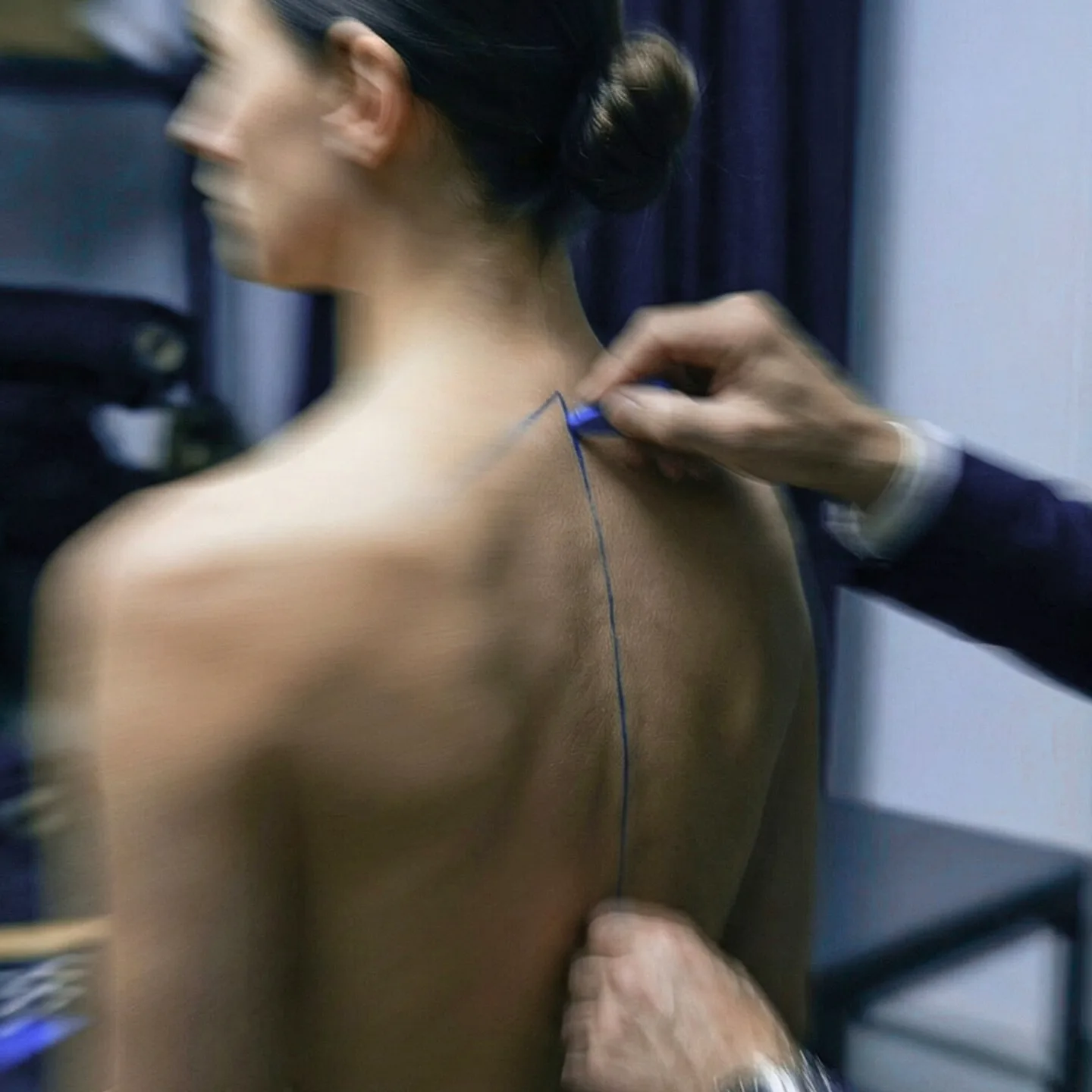 A fitting in progress: a line of blue thread run down the spine of a bare back, pinned at the shoulder by one hand and held at the waist by another, the room blurred around the movement.