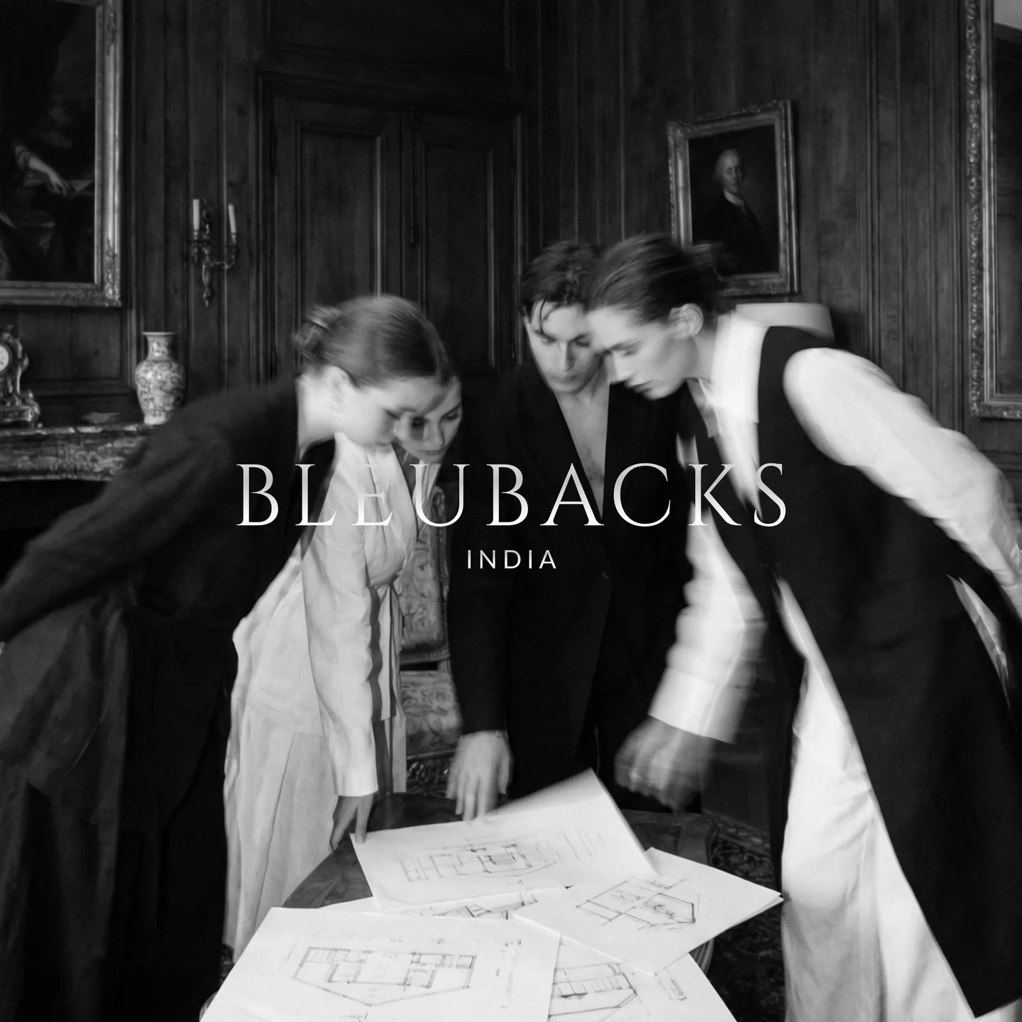 The Bleubacks India lockup in white over a black and white photograph of four people leaning over architectural drawings spread across a table in a panelled room.