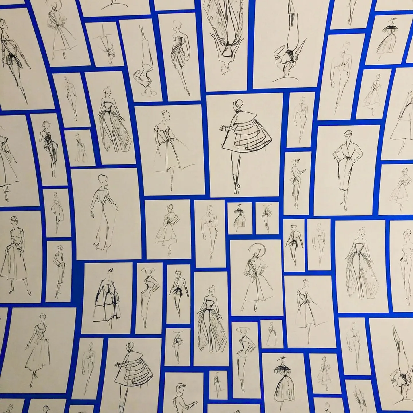 A wall of ink fashion sketches on cream cards, tiled edge to edge with lapis blue showing through the gaps between them.