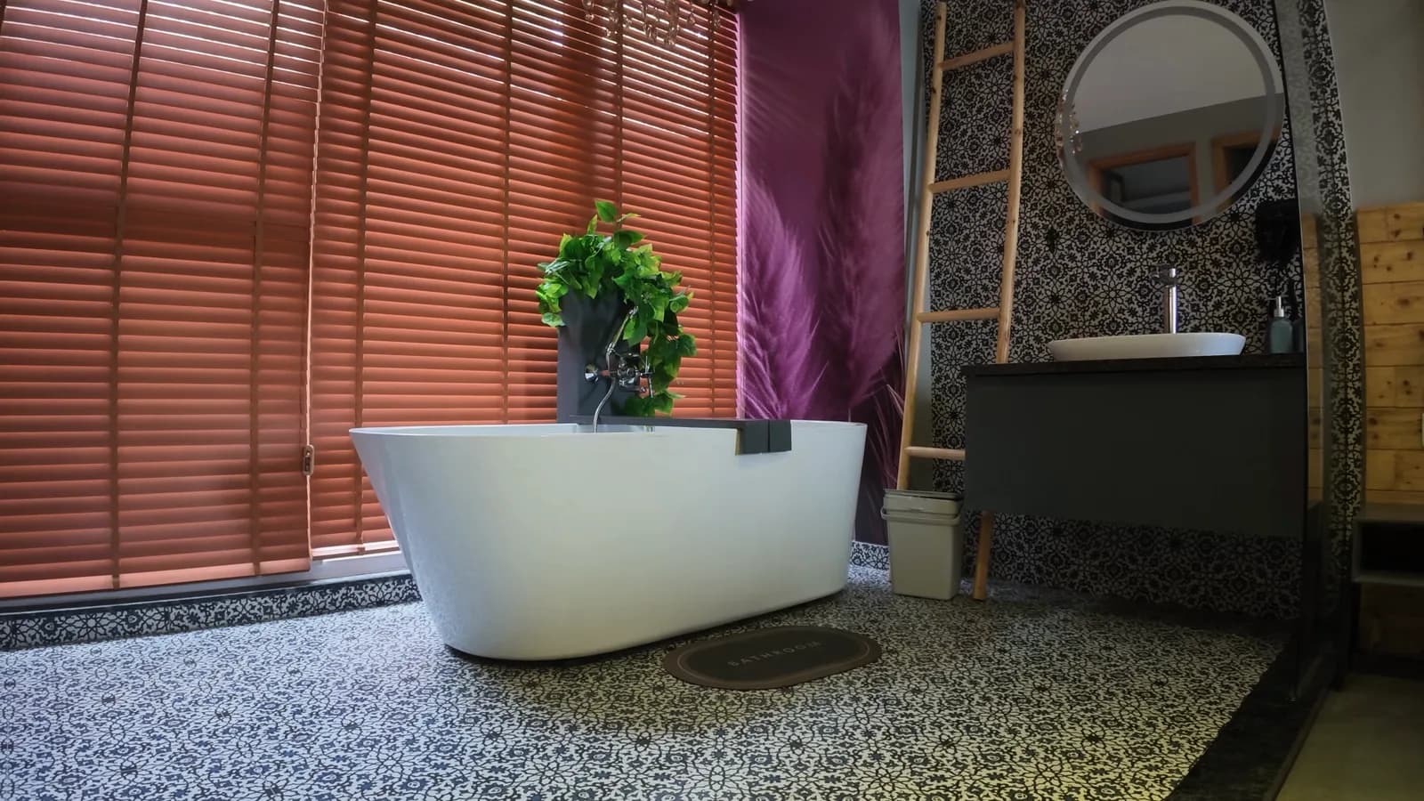 A freestanding bath on terrazzo beneath a wide window with timber blinds, a plant and a round mirror beside it.