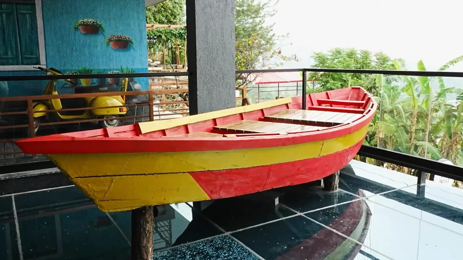 A red and yellow wooden boat set on a dark deck under a pergola, the hills falling away beyond the railing.