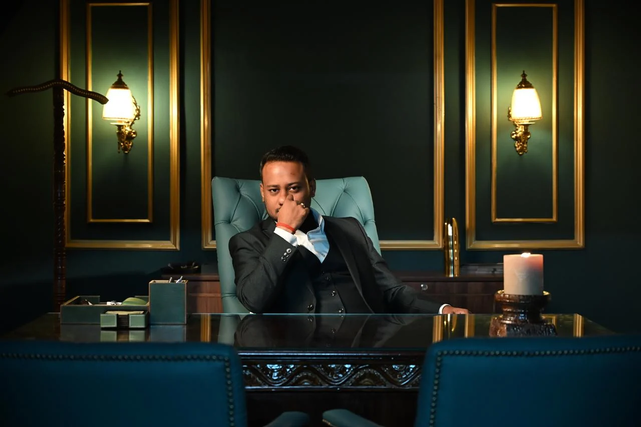 Pikova’s founder seated at a desk in a dark green panelled office, gold-framed wall lights on either side, a candle at one end of the glass top.